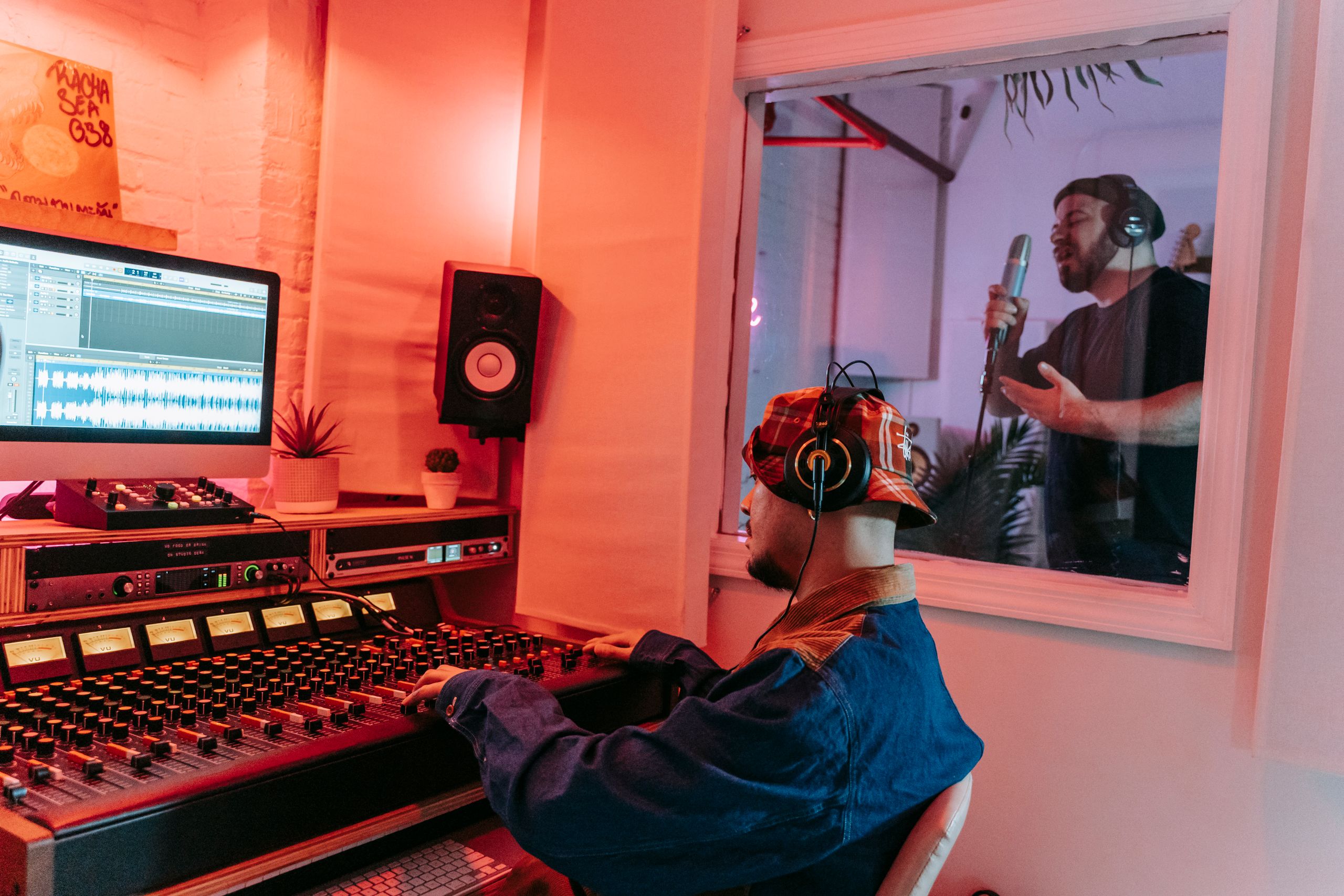 Music producer at work in a professional recording studio with a singer performing behind glass.